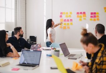 group of coworkers in a meeting room discussing a long list of sticky notes on the wall