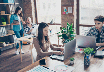 One female and one male administrative assistants sit at their desks typing on their laptops. Behind them, their boss discusses a report with another colleague