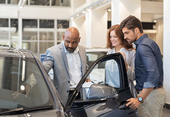 A balding salesman shows a car to a man and women in his showroom